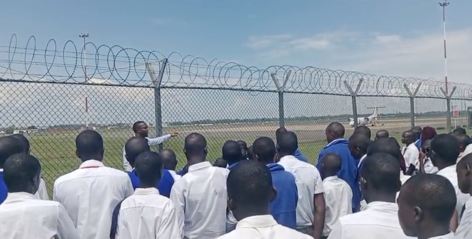 Students gathered at the airport perimeter fence while an airport staff member points out a plane on the runway