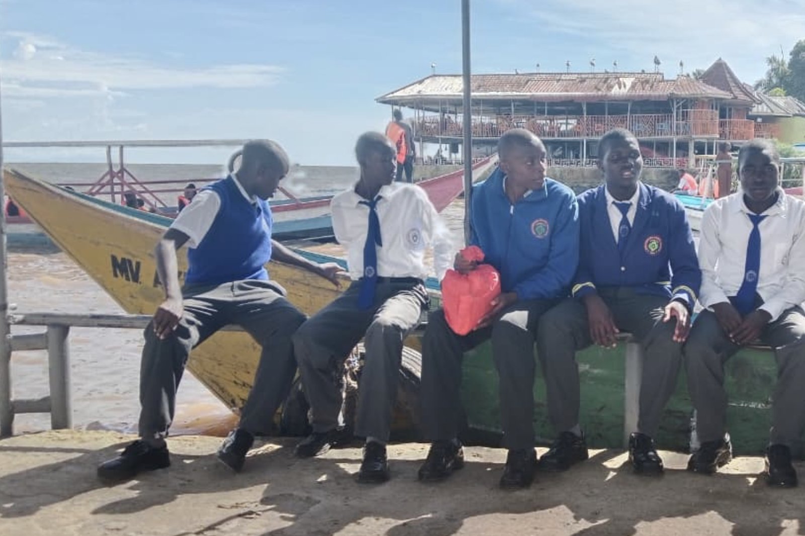 Five students sitting on the lakeshore in front of wooden boats on Lake Victoria